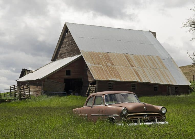 Vieille voiture américaine rouillée stationnée devant une grange, entourée d’herbes hautes, typique d’une découverte de grange.