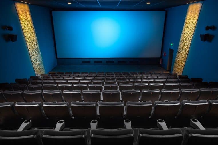 Salle de cinéma contemporaine aux sièges vides face à un grand écran, ambiance bleutée et murs texturés éclairés.