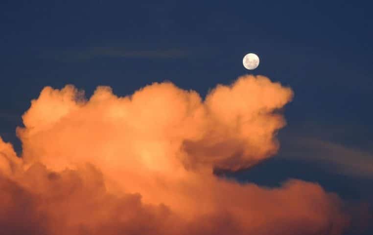 Disque de la pleine lune suspendu au-dessus d’un immense nuage orangé dans un ciel de nuit bleu profond