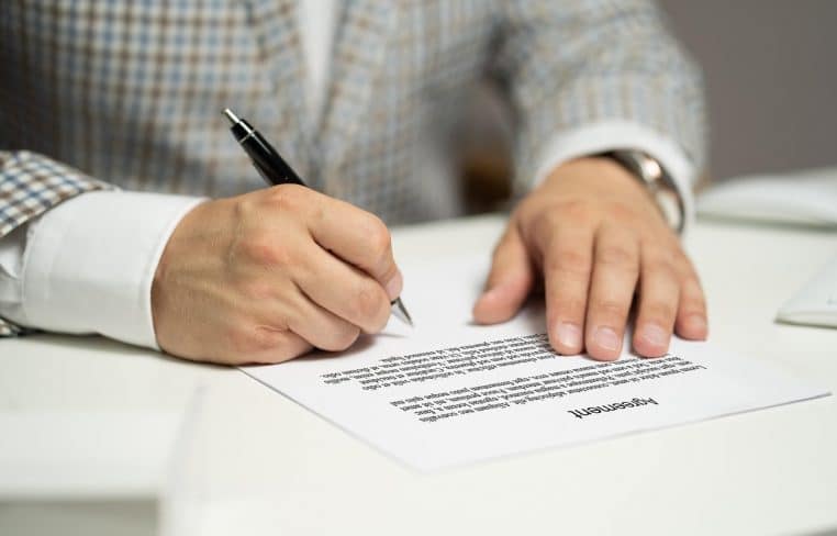 Plan rapproché sur un contrat en anglais avec la mention « Agreement », main masculine écrivant au stylo noir, veste à carreaux visible.