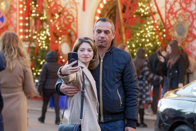 Couple prenant un selfie avec un smartphone devant des décorations de Noël lumineuses dans une rue animée