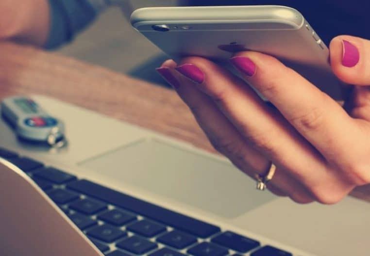 Gros plan sur une main tenant un smartphone au-dessus du clavier d’un ordinateur portable posé sur un bureau de travail