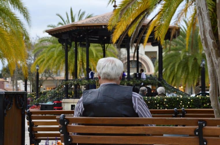 Homme senior de dos assis sur un banc dans un parc urbain bordé de palmiers, observant la vie autour de lui pendant sa retraite après le versement de sa pension.