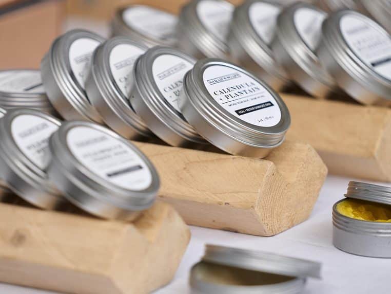 Femme absente, rangée de petits pots de crème pour le visage en métal posés sur un support en bois clair, évoquant un soin cutané artisanal.