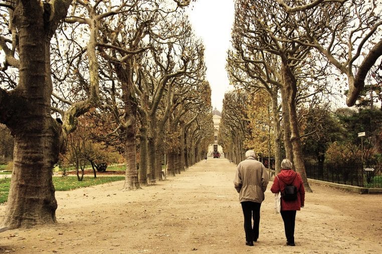 Couple de retraités marchant enlacés dans une rue de Paris à l’automne, feuilles au sol et façades haussmanniennes en arrière-plan.