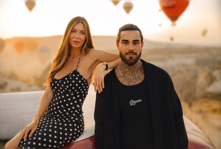 Un couple assis sur une voiture décapotable au coucher du soleil, avec des montgolfières floues en arrière-plan dans le ciel.