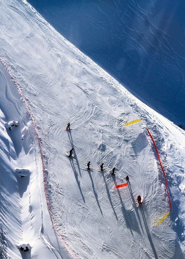Piste de ski inclinée vue en plongée, un groupe de skieurs descent la pente en file avec de longues ombres étirées sur la neige damée.