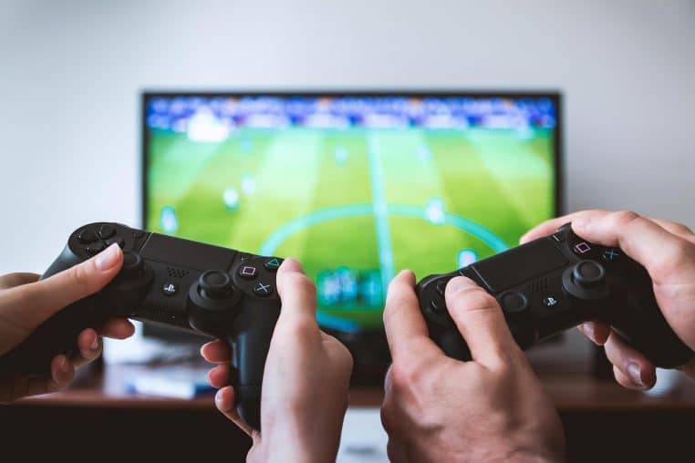 Deux joueurs assis dans un salon moderne manient des manettes de jeu devant une télévision allumée, illustrant la consommation électrique des consoles en veille.