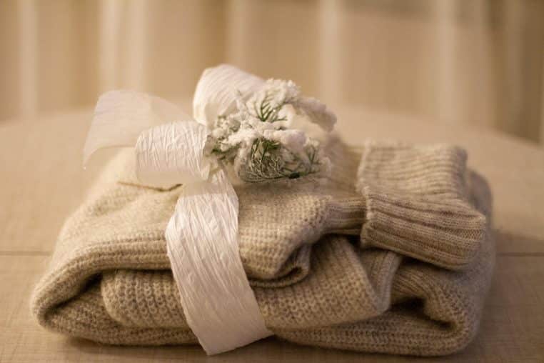 Pull en maille beige plié et noué d’un large ruban blanc avec un petit bouquet enneigé posé dessus, sur une table intérieure.