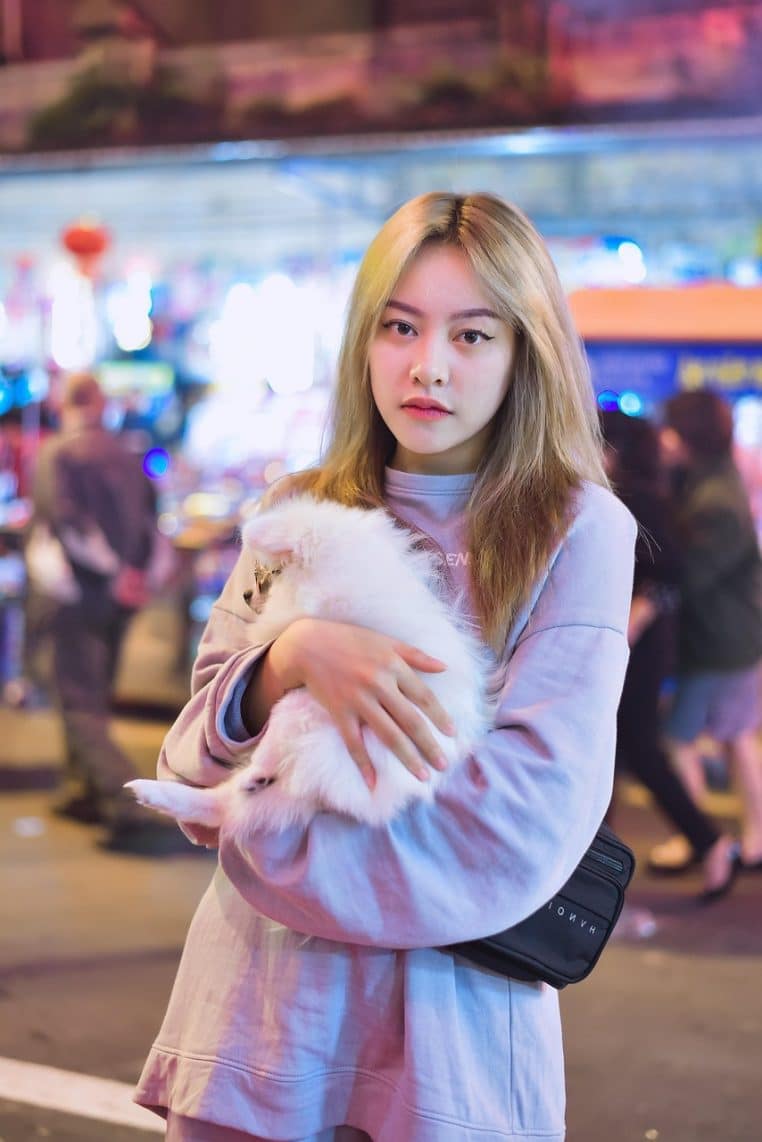 Jeune femme en ville serrant un petit chien blanc dans ses bras, éclairée par les lumières nocturnes, symbole de soutien affectif discret.