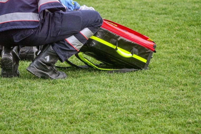 Sac d’intervention rouge posé sur une pelouse à côté d’un secouriste en uniforme, évoquant la rapidité d’action des équipes de secours.