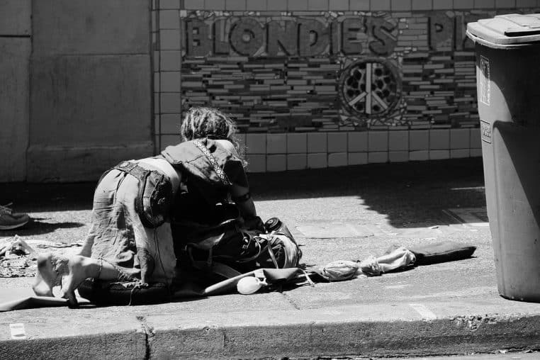 Femme assise sur le trottoir d’une grande ville, emmitouflée dans un manteau, un gobelet posé devant elle pour demander de l’aide.