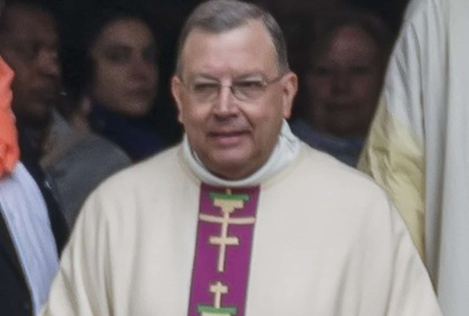 Gros plan centré sur le visage de Mgr Jean-Paul Gusching, en aube crème et étole violette, capté à la sortie d’une célébration.