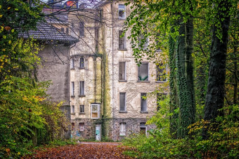 Façade d’une clinique abandonnée entourée de végétation, vitres cassées et murs marqués par le temps.