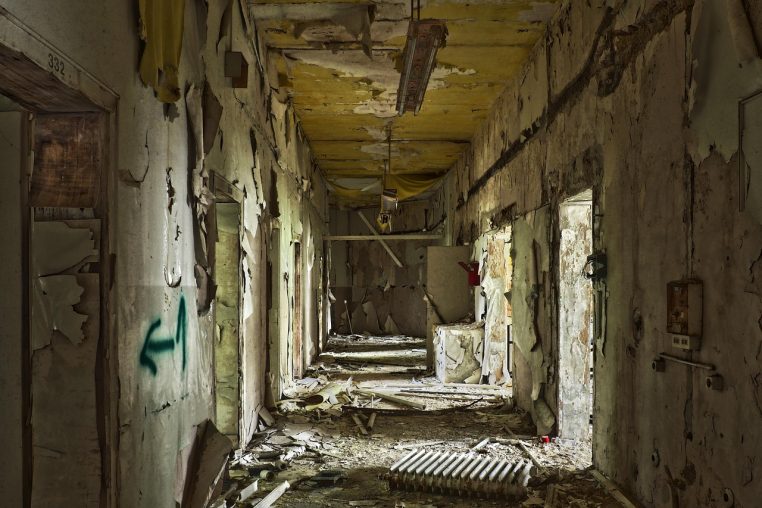 Salle d’un bâtiment abandonné, murs nus, grandes fenêtres et traces d’humidité, évoquant une ancienne zone de soins hospitaliers.
