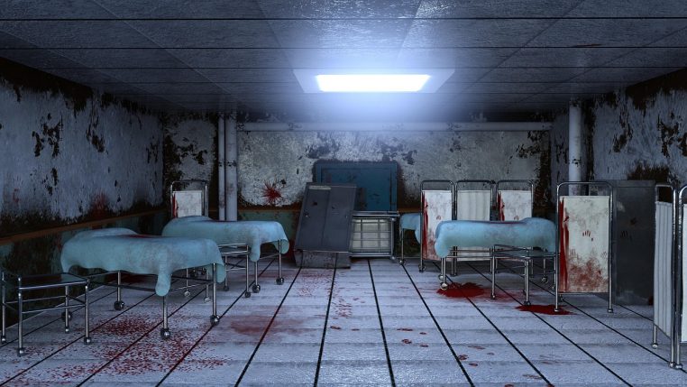 Chambre d’hôpital abandonnée avec plusieurs lits métalliques vides, murs décrépis et lumière froide entrant par les fenêtres latérales.