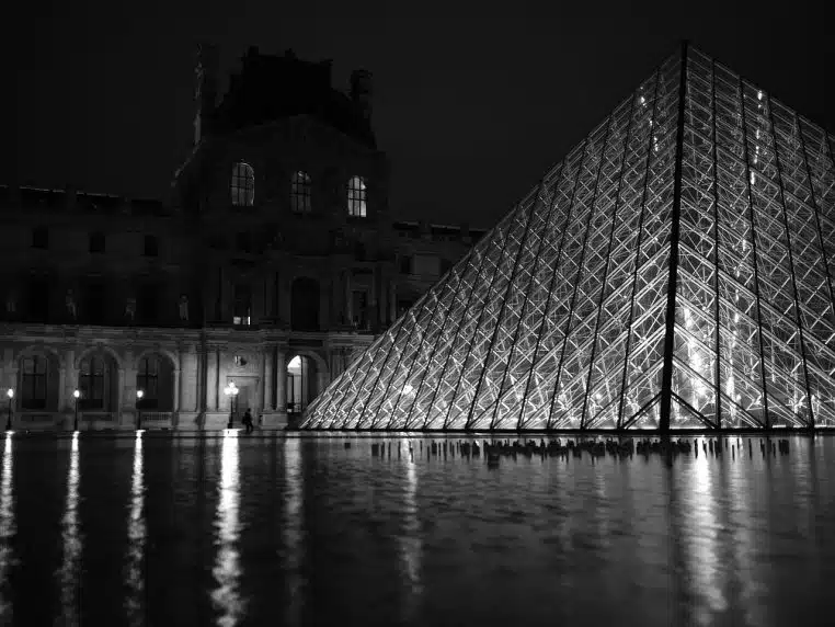 Vue nocturne du musée du Louvre et de sa pyramide de verre éclairée, reflétée dans le bassin sous le ciel sombre de Paris.