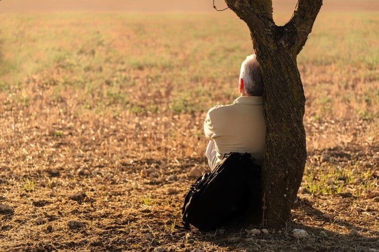 Homme assis seul contre un grand arbre au bord de l’eau, regard perdu au loin, dans un moment de réflexion silencieuse.