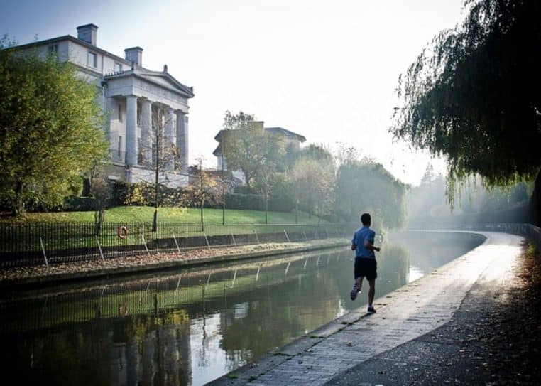 Homme courant au bord d’un canal au petit matin, dans un décor urbain calme, donnant une impression de rituel sportif immuable.