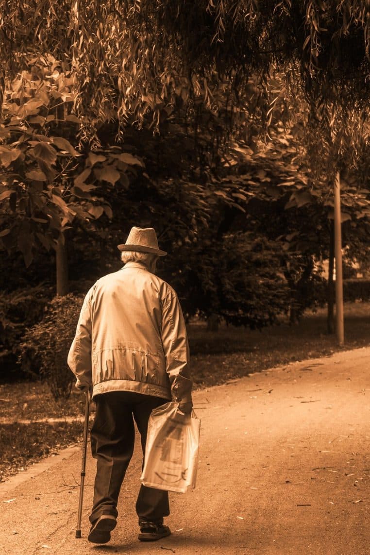 Homme âgé marchant avec un bâton le long d’une allée de parc bordée d’arbres, ambiance automnale et feuillage au sol en arrière-plan