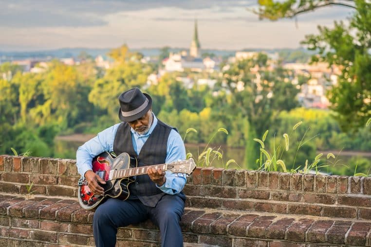 Homme senior assis sur un muret, jouant de la guitare électrique devant un paysage urbain flou, concentré et détendu au coucher du soleil.
