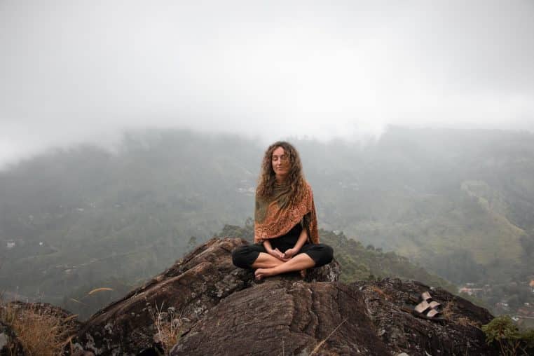 Personne assise en tailleur sur un rocher, les yeux fermés, méditant au milieu de montagnes couvertes de brume, atmosphère calme et silencieuse.