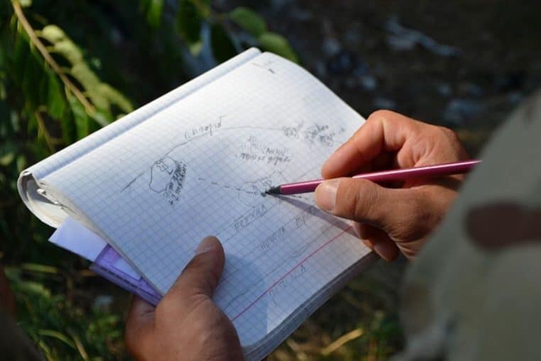 Plan serré sur des mains dessinant un schéma tactique au crayon sur un carnet quadrillé pendant une préparation de mission.