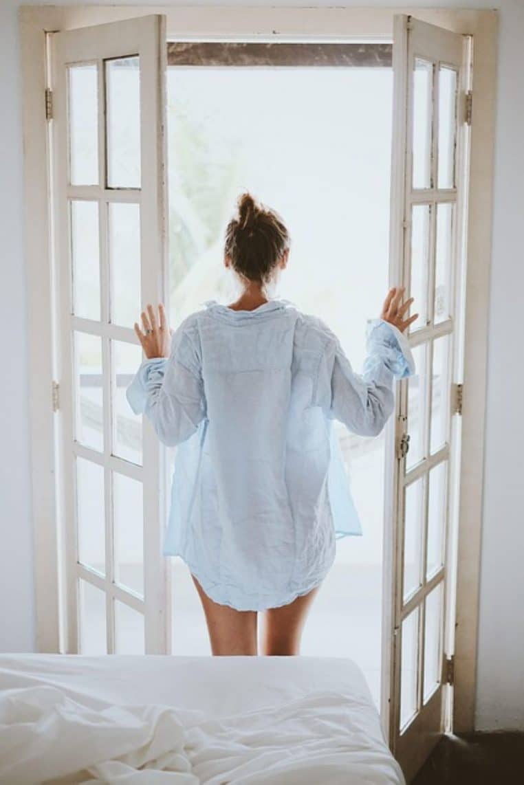 Jeune femme ouvrant les portes-fenêtres d’une chambre au petit matin, lit défait au premier plan et lumière blanche extérieure.