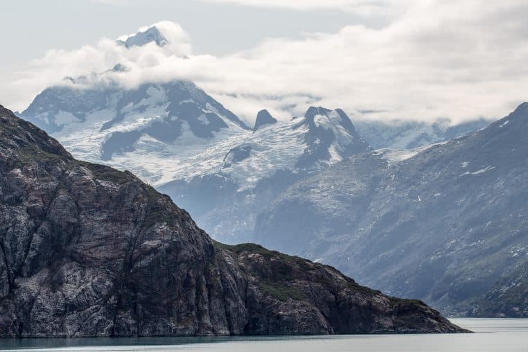 Chaîne de montagnes enneigées se reflétant dans un fjord calme sous un ciel nuageux, paysage typique des côtes sauvages d’Alaska.