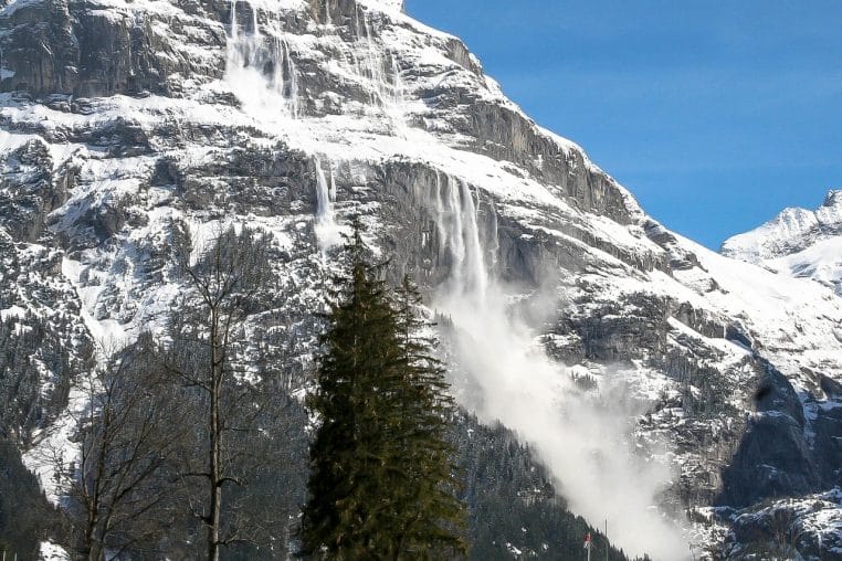 Coulée de neige descendant d’une paroi rocheuse dans les Alpes, montrant une avalanche en journée sous un ciel bleu parfaitement dégagé.