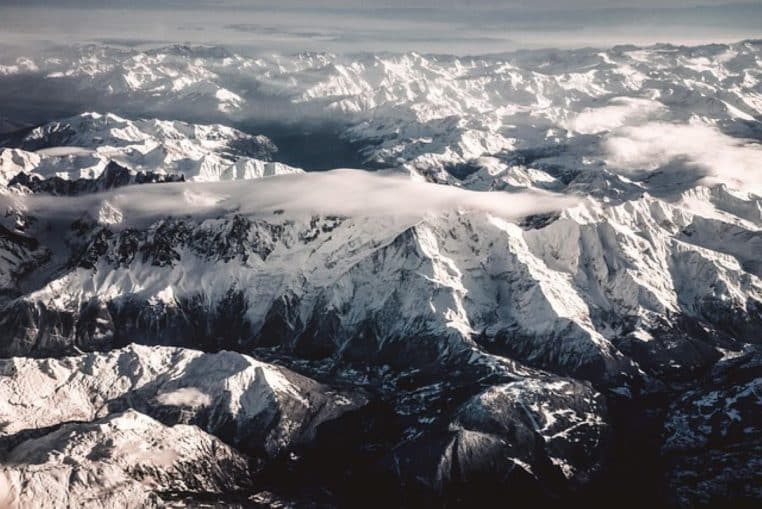 Vue aérienne d’une chaîne alpine entièrement recouverte de neige, avec un épais nuage accroché aux crêtes centrales du massif.