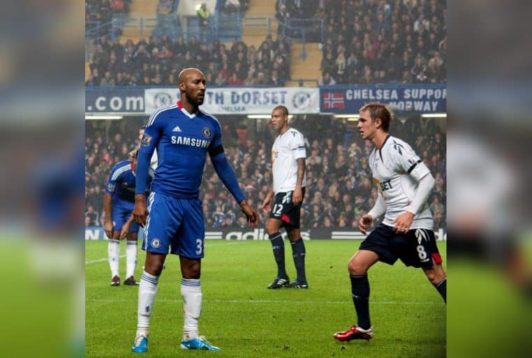 Nicolas Anelka en maillot de Chelsea durant un match face à Bolton, observant l’action au milieu des joueurs adverses.