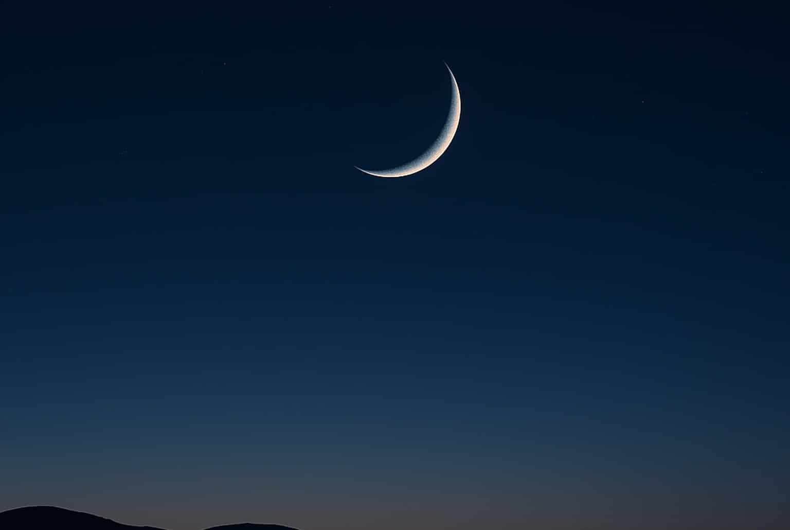 Fin croissant de Lune sur horizon montagneux et plan d’eau, ciel bleu nuit dégradé, calme annonçant la Nouvelle Lune.