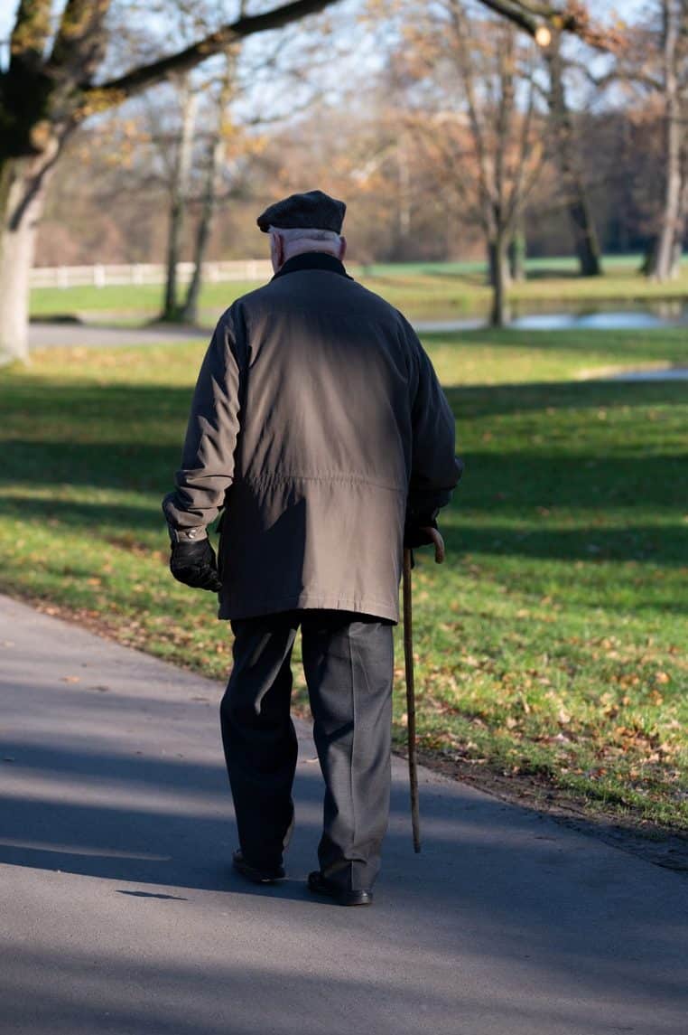 Homme âgé marchant avec une canne le long d’une petite route de parc, paysage naturel calme et lumière douce de fin de journée