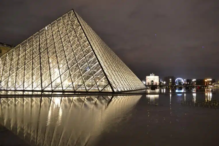 Pyramide du Louvre et bâtiments historiques environnants illuminés la nuit, sous un ciel profond au cœur de Paris.