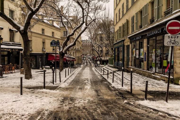 Rue parisienne bordée d’immeubles et de cafés sous la neige, chaussée partiellement déneigée et trottoirs encore recouverts.