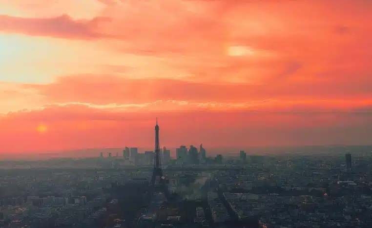 Vue panoramique de Paris au coucher du soleil, avec toits, gratte-ciel et tour Eiffel se découpant sur un ciel orangé.