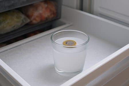 Gros plan d’un tiroir de congélateur ouvert, avec un verre glacé et une pièce d’un euro posée au-dessus de la glace.