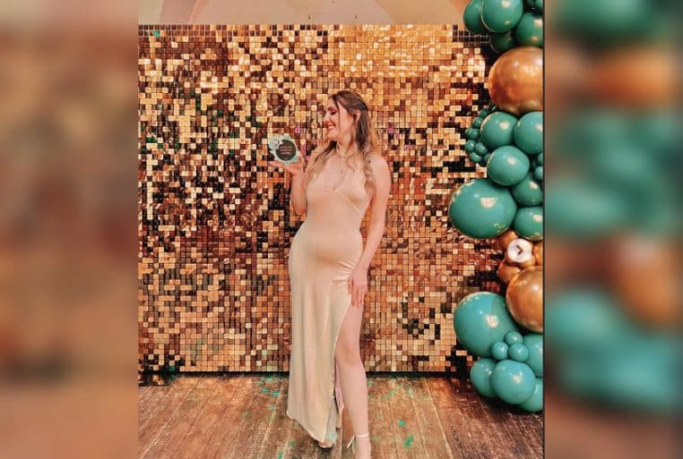 Jeune femme en longue robe champagne posant devant un mur pailleté doré, trophée en main, lors d’une soirée festive.