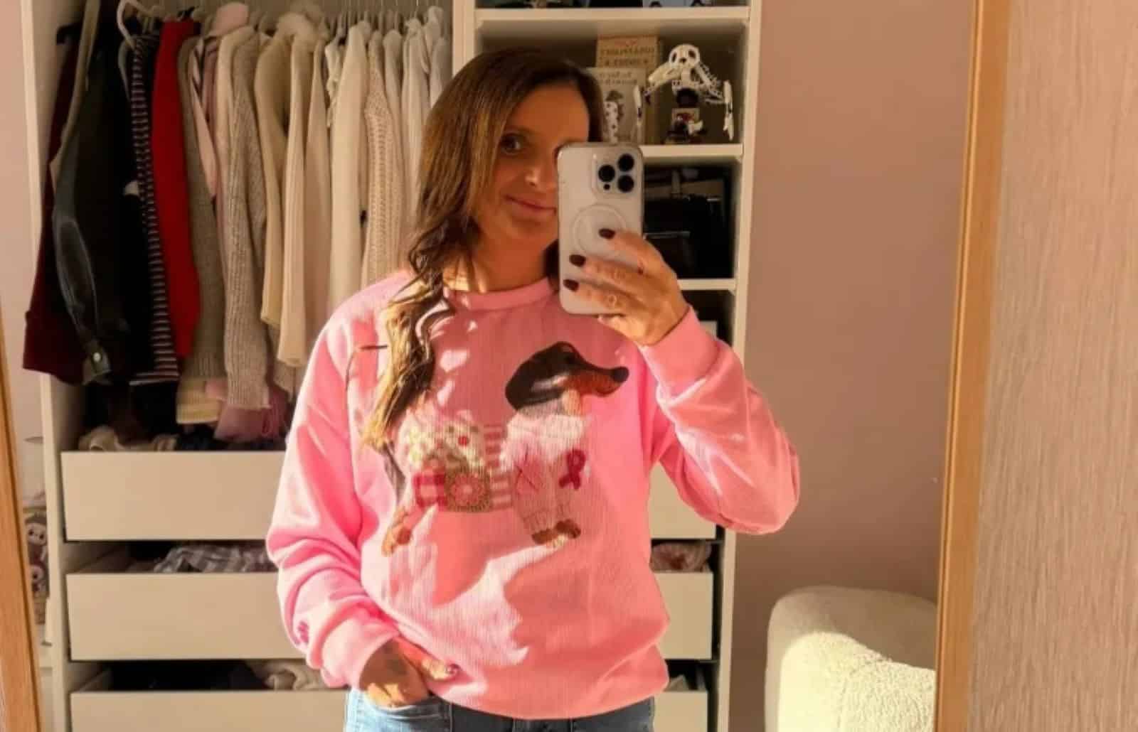 Selfie miroir de Sue Radford portant un pull rose à motif dachshund et ruban rose, devant un dressing baigné de soleil.