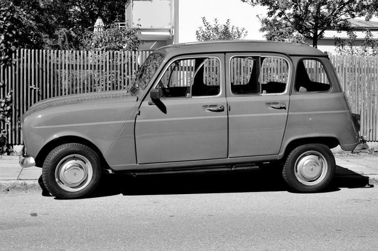 Renault 4L beige garée en noir et blanc le long d’une rue résidentielle, vue de profil, illustrant une voiture populaire des années 1970.