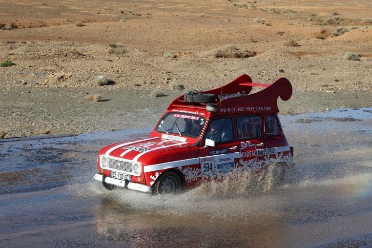 Renault 4L rouge engagée dans le 4L Trophy, roulant dans un gué au milieu d’un paysage désertique, couverte de stickers de sponsors.