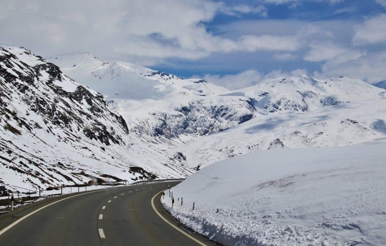 Route de montagne dégagée serpentant entre des pentes couvertes de neige fraîche sous un ciel d’hiver lumineux, symbole des conditions de circulation en altitude