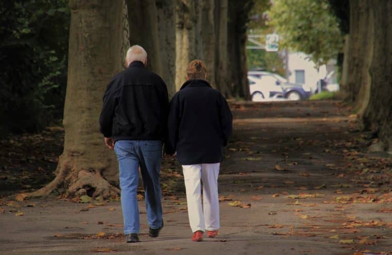 Deux personnes âgées marchent côte à côte dans une allée d’automne, entourées d’arbres dorés, concentrées sur leur échange.
