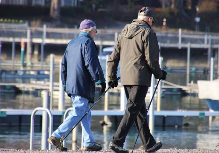 Couple de personnes âgées marchant côte à côte sur un chemin près de l’eau avec des bâtons de marche, ciel dégagé et décor naturel apaisant