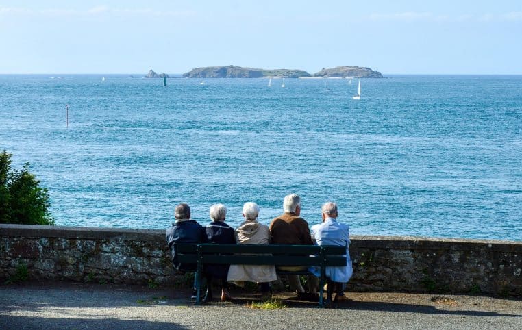 Groupe de quatre seniors assis sur un banc face à la mer, regardant des voiliers au large sous un ciel dégagé, moment calme de contemplation partagée.