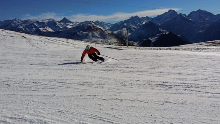 Skieur en combinaison rouge taillant une courbe sur une large piste des Alpes, avec une chaîne de montagnes enneigées en arrière-plan.