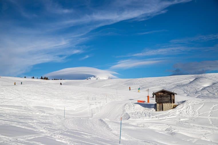 Station de ski française baignée de soleil avec une vaste piste blanche, une petite cabane en bois et plusieurs skieurs éparpillés sur la neige.