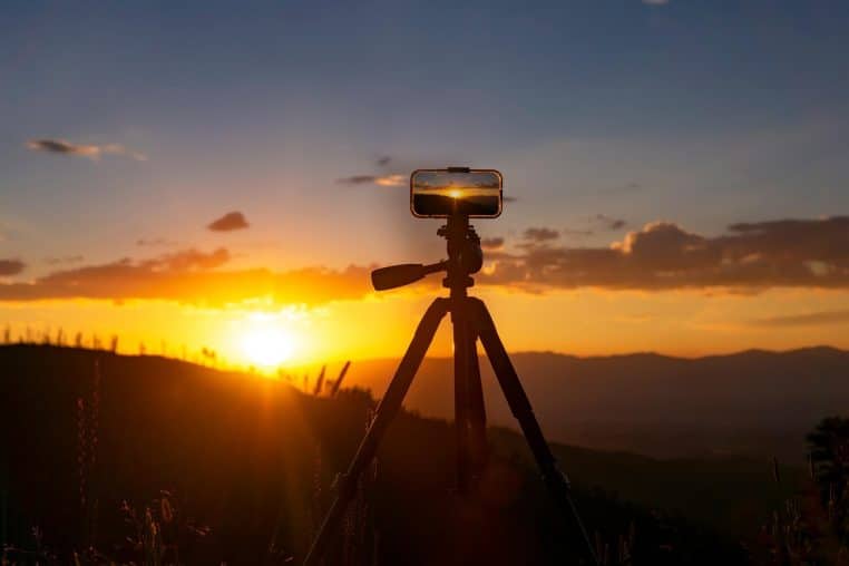 Smartphone fixé sur un trépied filmant un coucher de soleil orangé au-dessus des montagnes, illustrant un tournage de contenu en extérieur