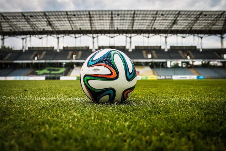 Un ballon de football posé sur une pelouse verte impeccable dans un grand stade éclairé, avec des tribunes floues en arrière-plan prêtes à accueillir les supporters.